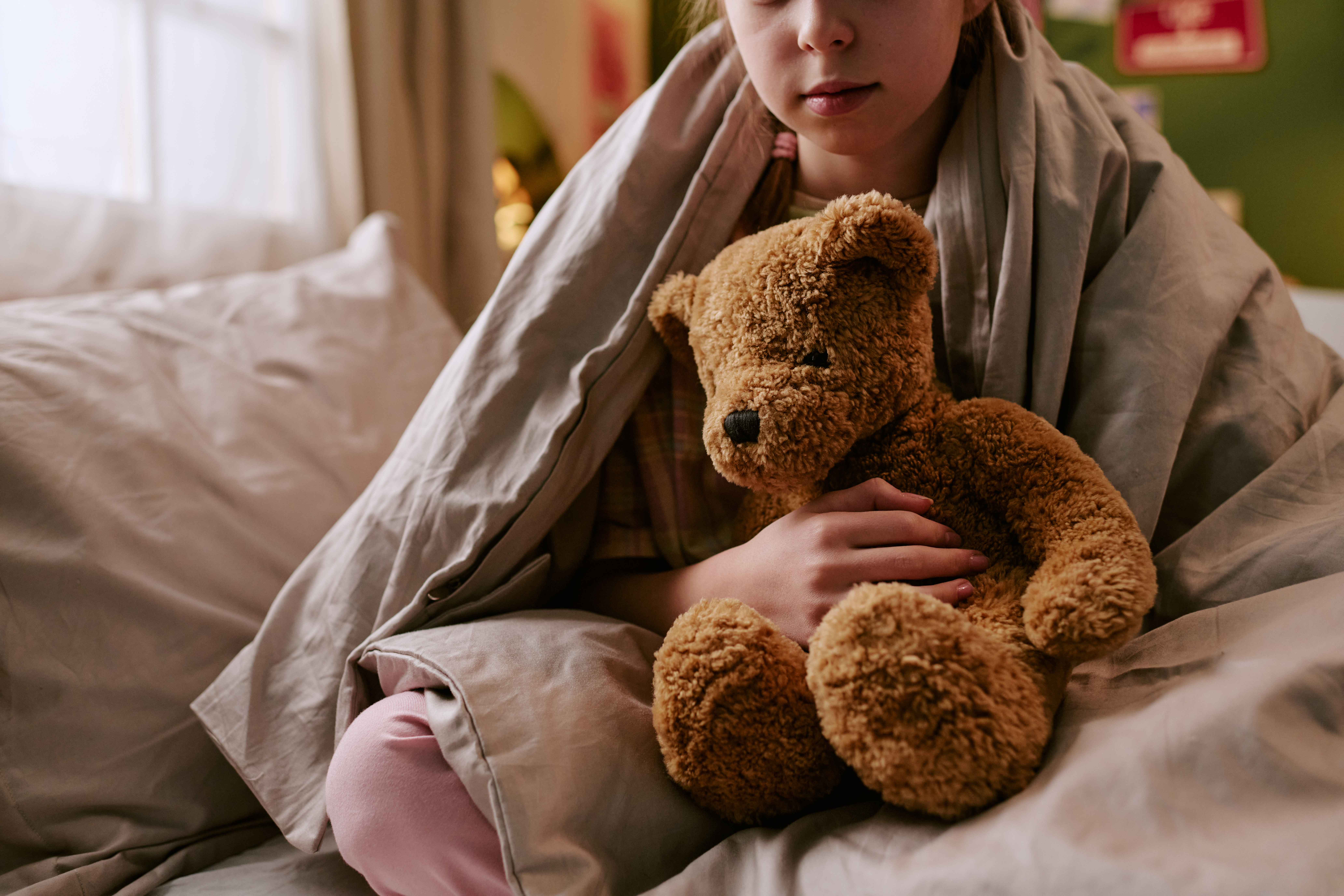 Caucasian child sitting on bed holding toy bear, partially covered with blanket, looking down with neutral expression, soft natural light illuminating face and hands