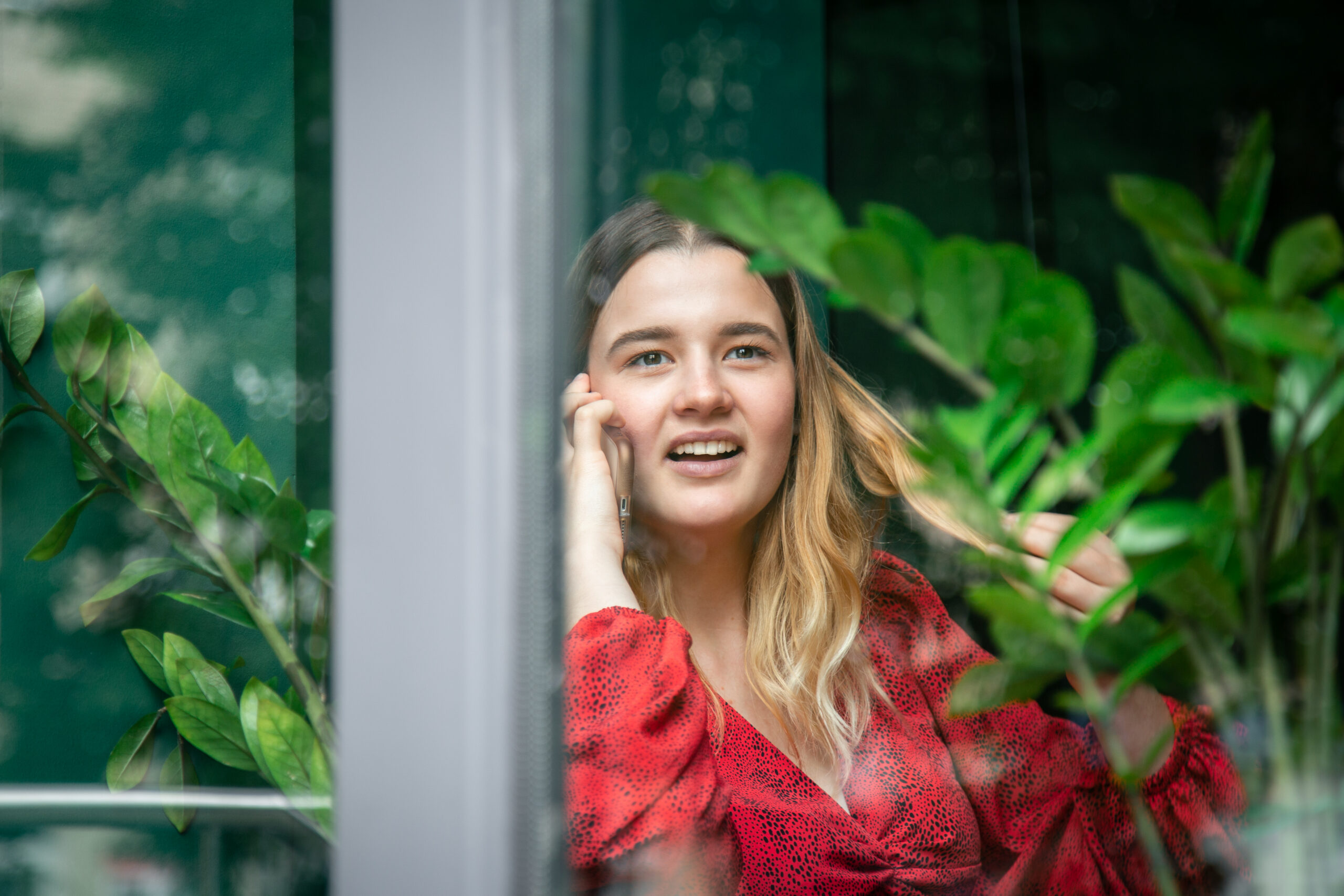 A girl in a red dress is talking on the phone in a cafe, the view from the street through the window.